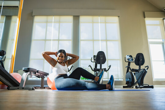 Plump young black woman exercising in gym lying on yoga mat doing leg raising and twisting exercises. Young attractive plus size woman doing abs workout. 