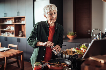 Focused senior woman seasoning her meal while cooking in a modern kitchen