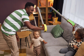 Side view portrait of happy Black father playing pirates with two children at home and holding toy swords