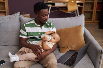 Portrait of young Black father bottle feeding cute baby boy while working from home with laptop