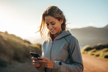 Young athletic Hispanic woman in sportswear with smartphone while training outdoor. Slender swarthy girl resting after jogging or workout in beautiful mountains. Active lifestyle for all ages. - obrazy, fototapety, plakaty