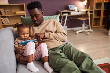 Portrait of young Black father teaching little girl using smartphone while sitting together on couch at home, copy space