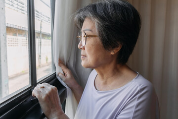 Asian old woman sitting on chair and opening white curtain while looking out the window, missing and waiting family and thinking something sad, living alone. - obrazy, fototapety, plakaty