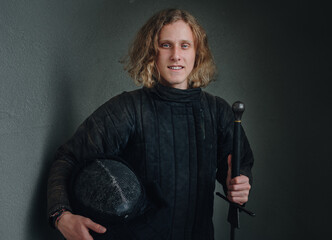 Portrait of a smiling long-haired young teen dressed in black historical fencing armor with a long medieval sharp sword