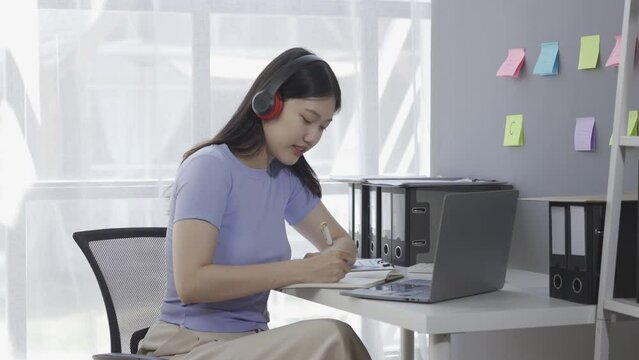 Young Asian female student wearing headphones surfs the internet on a laptop while taking an online class or lecture and doing a Google Zoom video call.