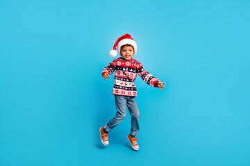 Full size photo of satisfied kid wear red pullover jeans santa hat jumping having fun on christmas party isolated on blue color background