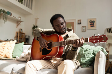Front view portrait of young Black man playing acoustic guitar while enjoying free time in cozy home, copy space - obrazy, fototapety, plakaty