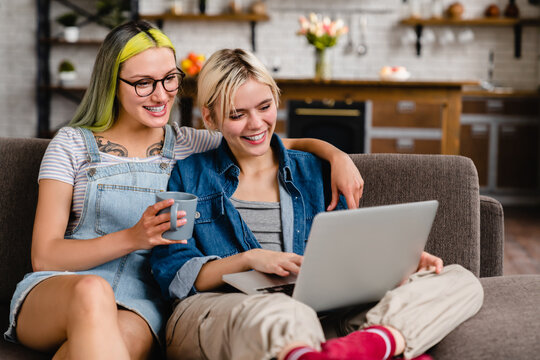 Happy cheerful young sisters girlfriends lgbtq lesbian couple chilling on the sofa, hugging cuddling and watching movie series film webinar on laptop together at home online