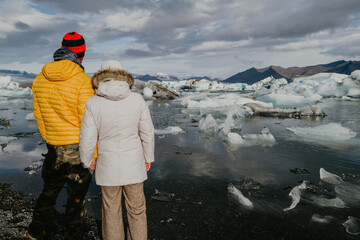 Couple in winter clothes standing overlooking Jökulsárlón glacier lagoon in Iceland - obrazy, fototapety, plakaty