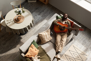 Top down view of Black young man playing acoustic guitar lying on sofa in cozy home lit by...