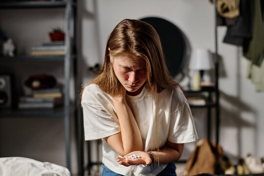Desperate teenager in white t-shirt holding pills on palm and looking at them while sitting in front of camera and trying to commit suicide