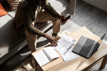 High angle portrait of Black adult man doing taxes at home in sunlight with financial documents on...