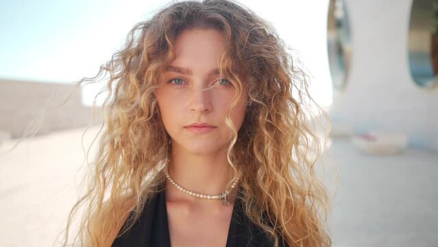 Close up portrait of woman with curly hair posing outside. Relaxed blonde lady with hair gently blown by wind standing outdoors in city