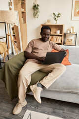 Vertical full length portrait of young African American man using laptop at home and relaxing in...