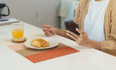 Happy beautiful Asian woman with phone eating fresh salad at table in kitchen and using application texting on smartphone while eating healthy food.