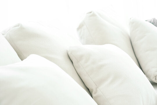 Closeup image of a pile of soft pillows made from white fabric used to decorate a sofa in a living room. There is natural light. It shines brightly from behind.