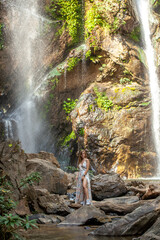 Young woman on rock under massive waterfall in Thailand. Experience the power of cascading water. Woman in dress at waterfall. Explore the jungles for an unforgettable travel experience.