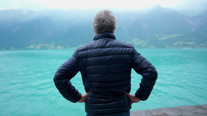 Back mature man standing in drizzle rain while staring at scenic view of lake and mountains,...