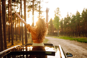 A young woman feels freedom leaning out of a car window in a sunny forest. A traveler enjoys a sunny day from the car window. Travel concept. Active lifestyle.