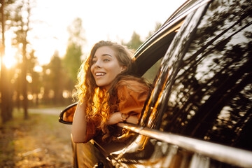 A young woman feels freedom leaning out of a car window in a sunny forest. A traveler enjoys a...