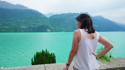 Pensive woman standing by natural lake view with mountains in background. Contemplative female person looking at lakeshire, enjoying nature's calm and serenity