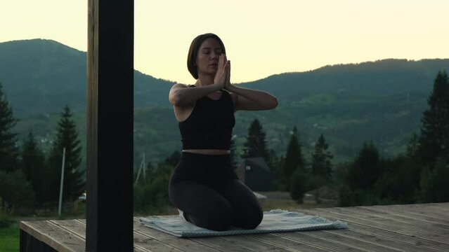 Young woman doing yoga on wooden terrace with mountains view. Female silhouette meditating and practicing yoga poses at dusk. Nature landscape