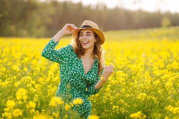 A happy woman in a hat walks through a blooming rapeseed field. Beautiful woman posing in a rapeseed field on a sunny day. Concept of nature, relaxation.