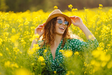 Naklejka na ściany i meble A happy woman in a hat walks through a blooming rapeseed field. Beautiful woman posing in a rapeseed field on a sunny day. Concept of nature, relaxation.
