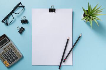 Top view of blank notebook, glasses, pen, calculator and green plant on blue office desk