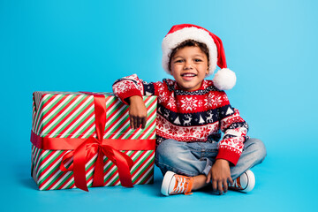 Obraz premium Portrait of satisfied little kid with wavy hair wear red pullover santa hat sit hold arm on present isolated on blue color background