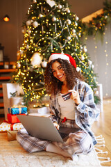 Fototapeta na wymiar Smiling woman with curly hair with a credit card, gifts and a laptop. A young woman at home near the Christmas tree makes online purchases. Shopping concept, holiday.