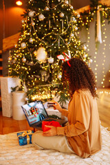 Smiling woman with curly hair with a credit card, gifts and a laptop. A young woman at home near the Christmas tree makes online purchases. Shopping concept, holiday.