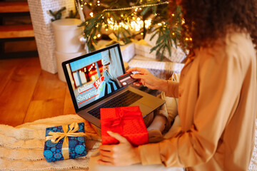 Naklejka na ściany i meble Smiling woman with curly hair with a credit card, gifts and a laptop. A young woman at home near the Christmas tree makes online purchases. Shopping concept, holiday.