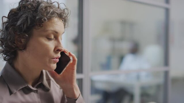 Close up of impatient Caucasian woman sitting in hospital hallway and speaking on phone while waiting for her turn
