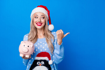 Photo portrait of attractive blonde woman point empty space piggy dressed penguin sweater x-mas hat isolated on blue color background