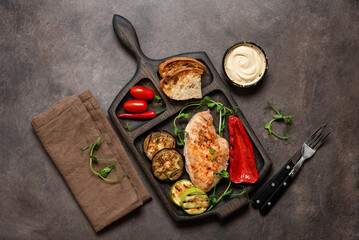 Grilled chicken fillet with grilled vegetables, sauce, toast on a wooden cutting board. Dark rustic background. Top view, flat lay, copy space