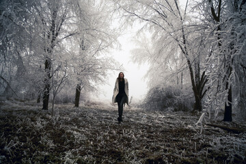 Woman in white coat walking through snowy forest on cold freezing foggy day.