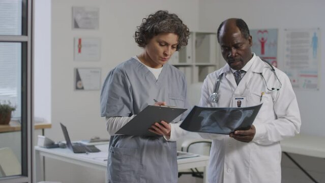 Medium shot of black male doctor analyzing chest x-ray scan and female Caucasian apprentice writing results while standing in clinic office