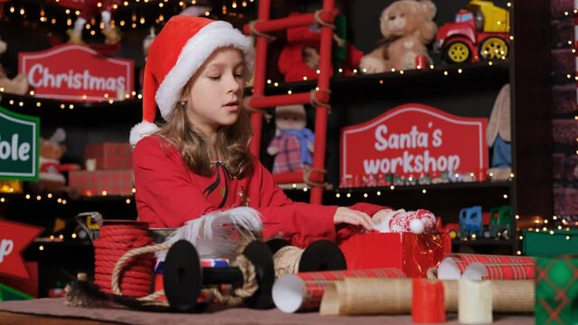 Little Santa helper packing gift at workshop, lots of toys and gift wrapping materials around her