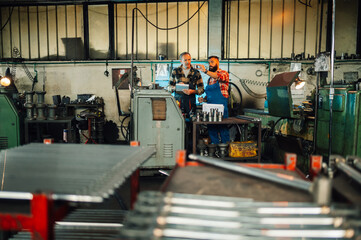 Wide shot of two metal turners looking at a metal piece by a lathe machine.