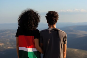 Rear view of a woman and a man standing on a mountain. The woman is wearing a T-shirt in the color of the Palestinian flag. You look at the endless landscapes of a country.