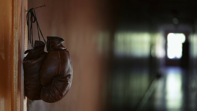 In the close up, shot, brown leather boxing gloves hang on a cord from a rusty nail on a door frame. In the background is an empty, dark corridor, someone has left their sport equipment