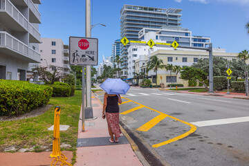 Beautiful view of woman walks down Collins Avenue on scorching, sunny day, shielding herself from...