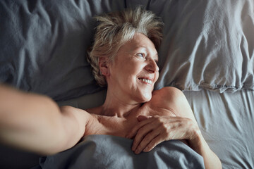 Portrait of a radiant mature woman taking a selfie while relaxing in bed