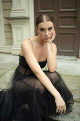 Fototapeta na wymiar a 30-year-old girl sits near the house in a black, transparent, revealing dress. Style and elegance, fatal girl, photo shoot in a black dress, cheating