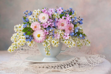 A bouquet of spring flowers: bird cherry, daisies, forget-me-nots in a cup on the table, a beautiful composition, still life, a holiday card. - obrazy, fototapety, plakaty