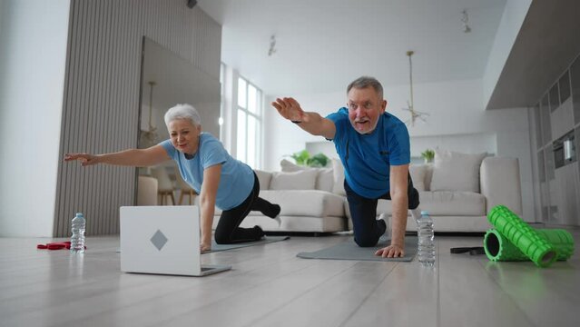 Happy senior couple doing pilates stretching exercise together synchronously at home on mats. Man woman training together online on laptop. Home fitness, stretching, slow training, sport concept.