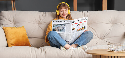 Young woman with headphones reading newspaper on sofa at home