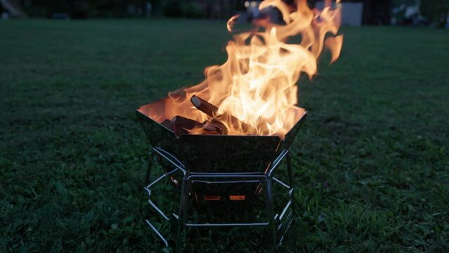 Flames rise from small portable grill on grassy field at dusk