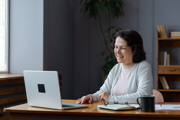 Happy middle aged senior woman sit with laptop talk on video call with friends family. Laughing mature old senior grandmother having fun talking speaking with grown up children online